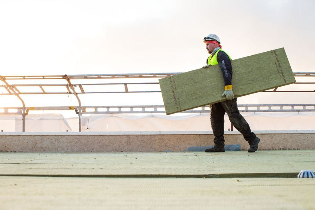 Arbeider i verneutstyr bærer en stor isolasjonsplate over en takflate.
