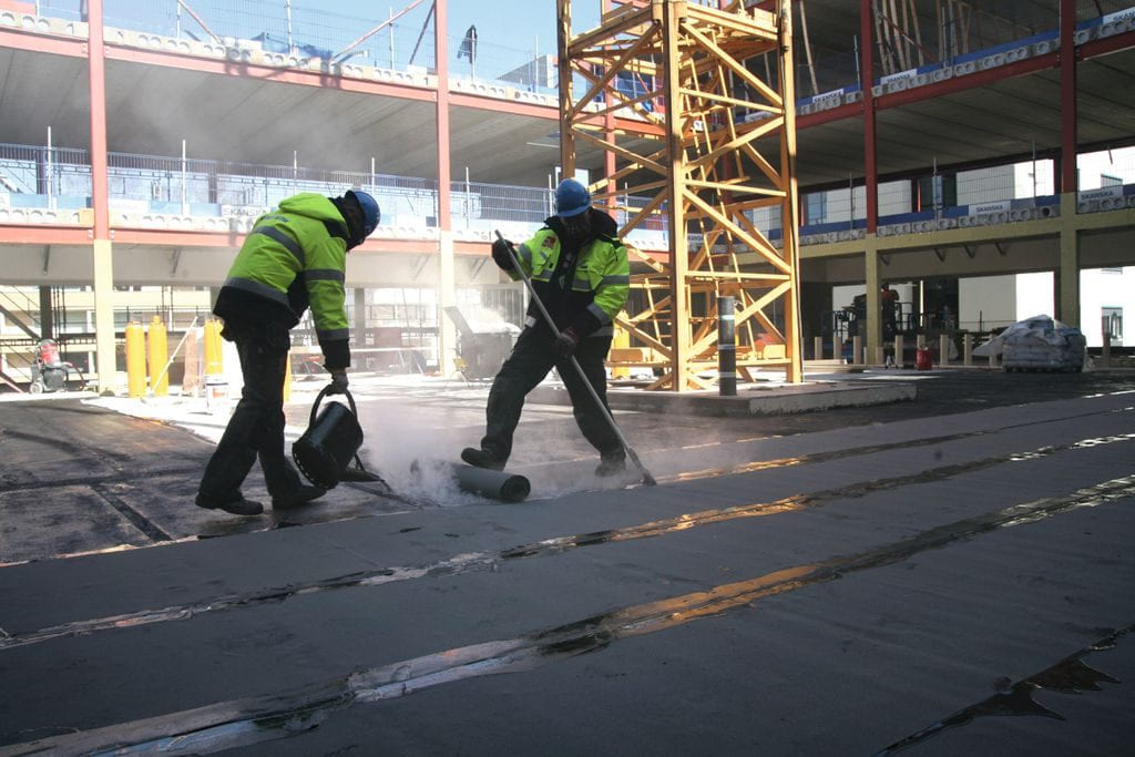 Hesselbergtaks fagarbeidere legger dobbel asfaltmembran med varm asfalt under grytetekking på byggeplass.