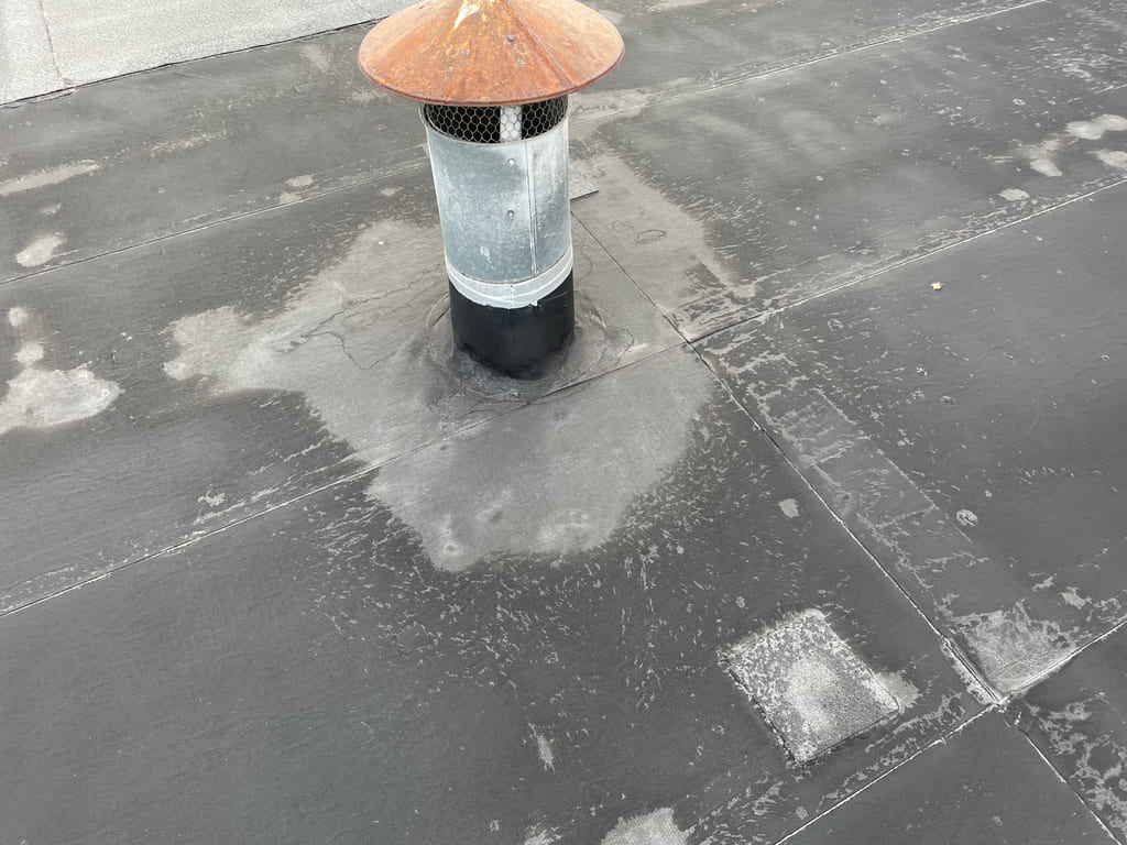 "Nærbilde av et takbelegg med tydelig slitasje rundt en gjennomføring med ventilasjonsrør. Overflaten er preget av misfarging, uttørking og nedbrutt sjikt – typiske tegn på manglende UV-beskyttelse over tid.