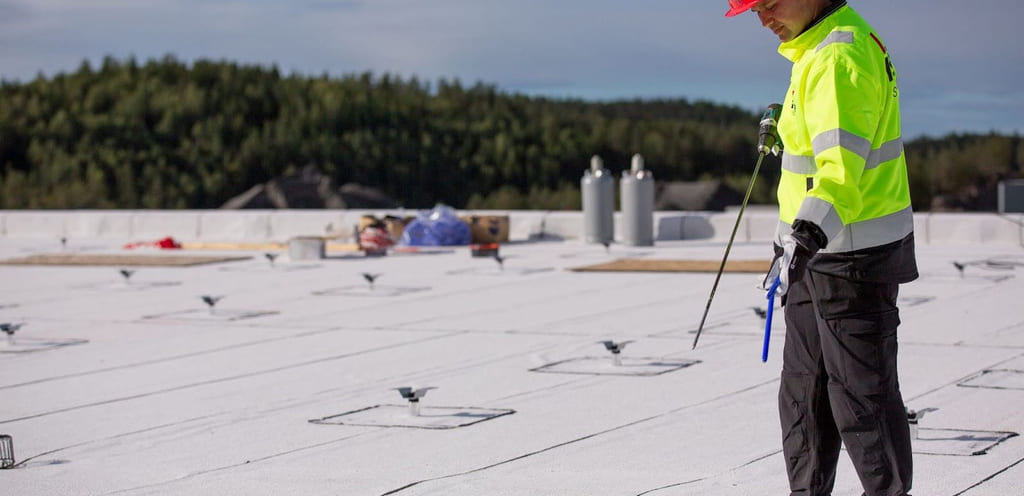 akarbeider i refleksjakke og rød hjelm kontrollerer festeanordninger på lyst takbelegg med spesialverktøy. Utsikt over skogkledd landskap i bakgrunnen, med utstyr og gassflasker synlig på taket.