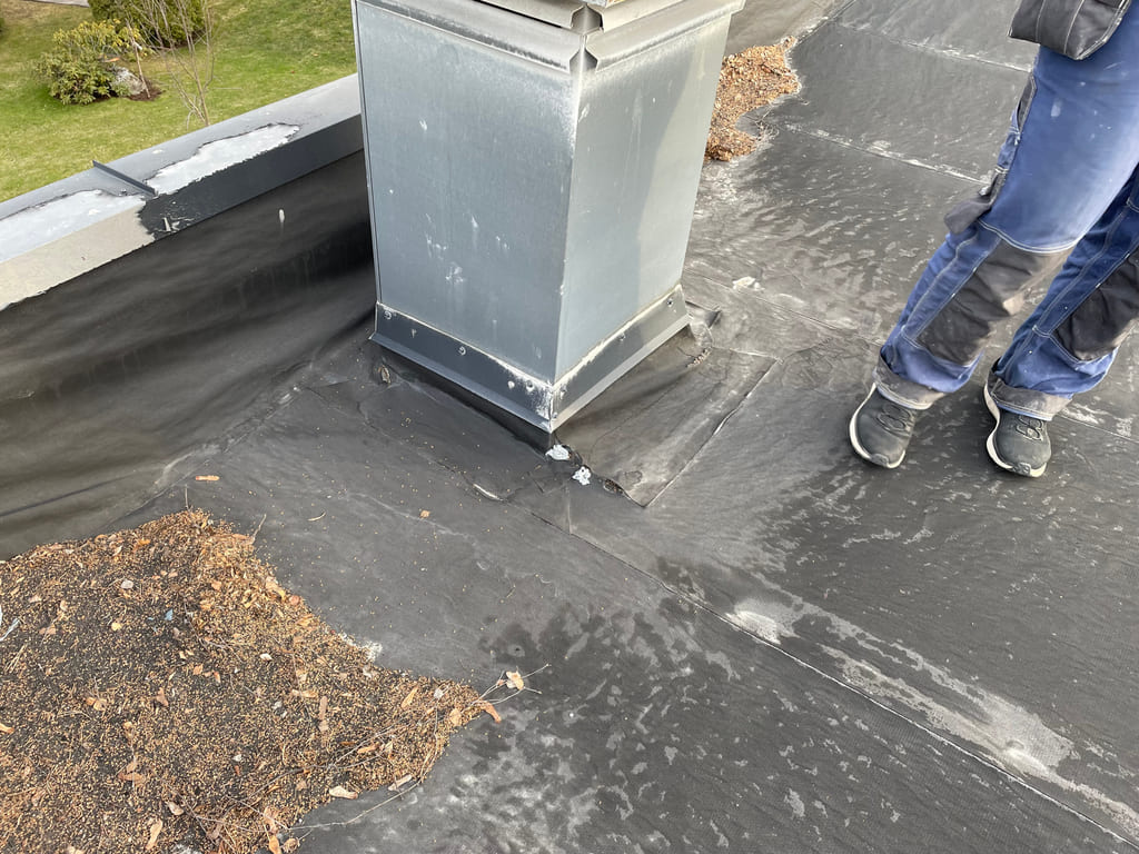 Nærbilde av eldre takmembran rundt en ventilasjonsgjennomføring, med synlige skjøter og ujevn tetting ved foten.