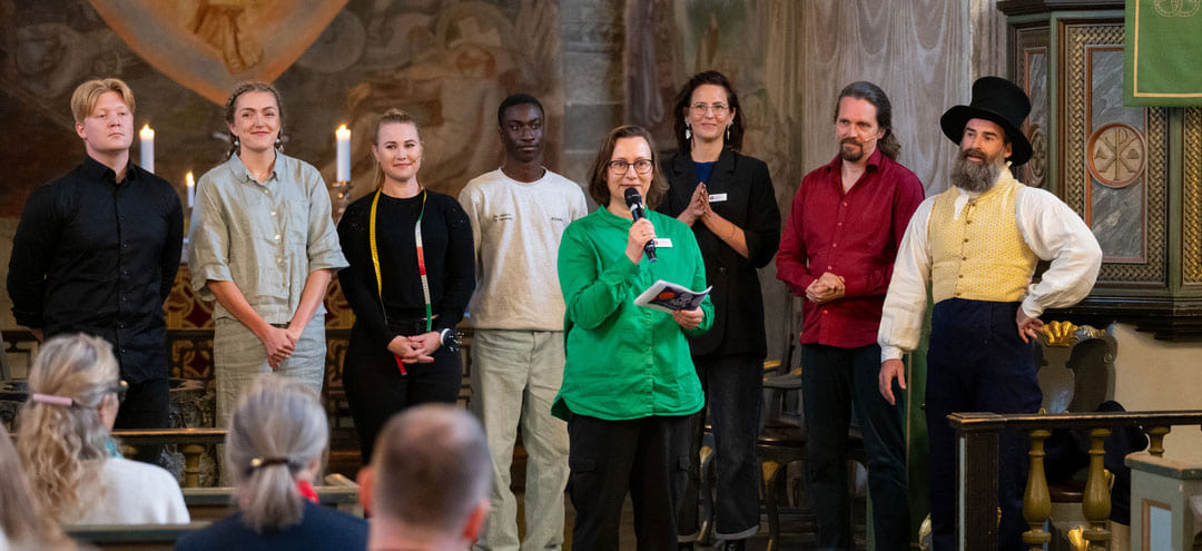 apning_av_SPOR_i_Stiklestad_kirke_KUL_20028_Foto_Trine_Ruud_Gronningen_crop3 Gruppe av forskjellige individer som står på scenen i en kirke, med en person i grønt som snakker inn i en mikrofon.