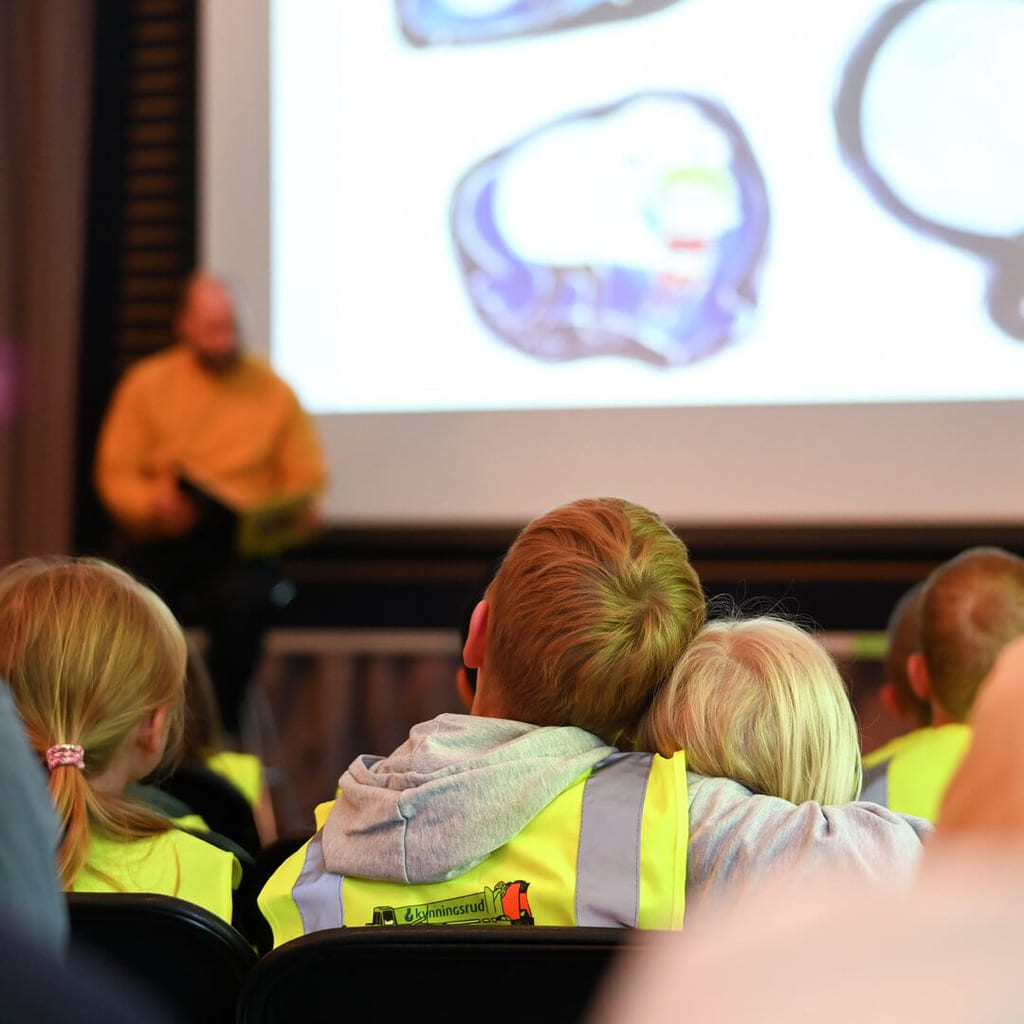 Barn med refleksvester ser på presentasjon mens de holder rundt hverandre.