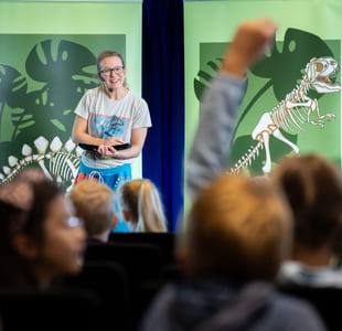 En kvinne står på en scene og snakker til en gruppe barn. Ett av barna rekker opp hånden.