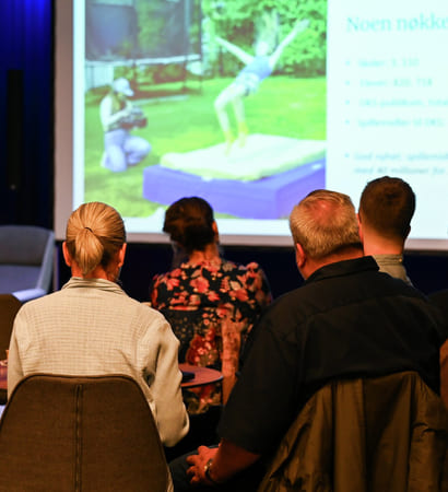 Publikum ser på presentasjonen med bilde av en person som hopper opp på en madrass utendørs. Projektorlerret og høyttaler er synlige.