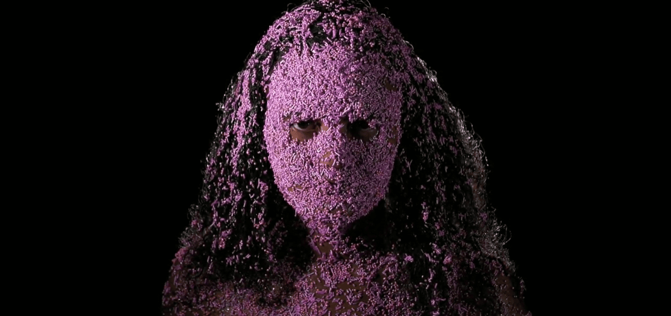 En kvinne står foran en sort bakgrunn og ser i kameraet med rosa kakedryss klistret i håret og ansiktet