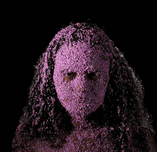 En kvinne står foran en sort bakgrunn og ser i kameraet med rosa kakedryss klistret i håret og ansiktet