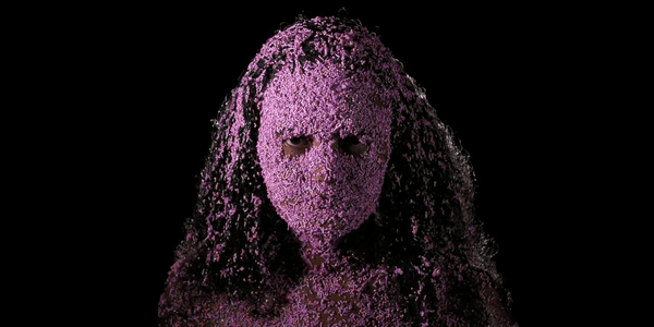 En kvinne står foran en sort bakgrunn og ser i kameraet med rosa kakedryss klistret i håret og ansiktet