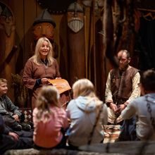 En kvinne og en mann i vikingklær sitter foran noen store trefigurer som forestiller vikinger. De smiler og forteller til en gruppe elever som sitter på gulvet med ryggen til kameraet