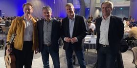 Fire menn står på rekke og smiler til kamera. Den ene holder en gitar.