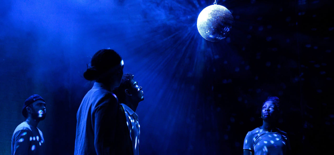 Fire skuespillere står på en blåbelyst og røykfylt scene. De ser opp på en discokule som henger i taket