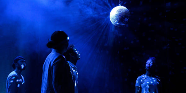 Fire skuespillere står på en blåbelyst og røykfylt scene. De ser opp på en discokule som henger i taket