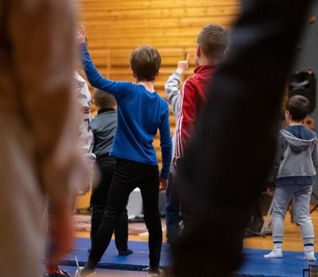 Barn i et gymsal rekker opp hendene nær en klatrevegg, omgitt av blå matter og trevegger.