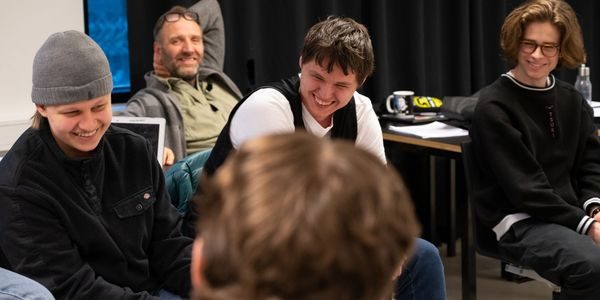 Elever sitter i en ring, en lærer sitter bak dem. De ser ned og smiler.