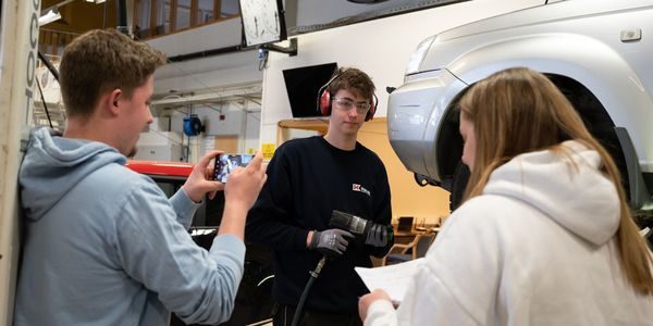 En elev filmer en annen mens en tredje elev ser ned i et ark