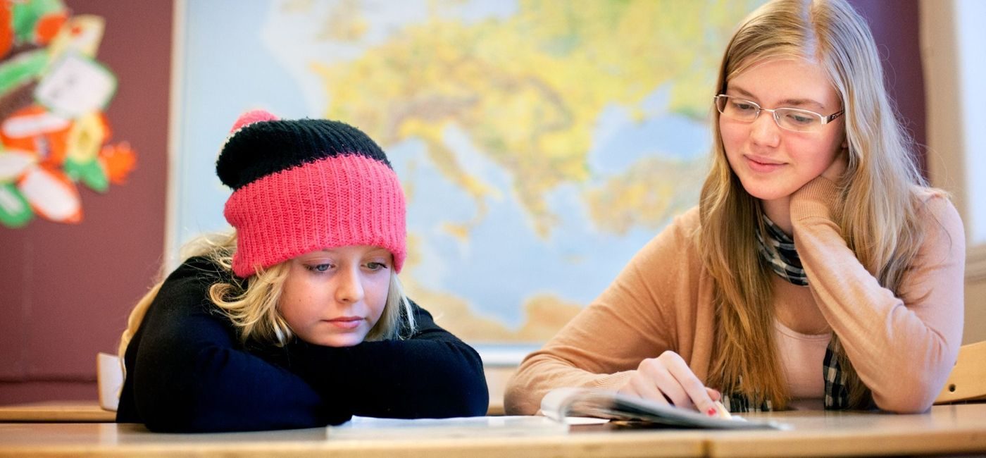 en elev med hatt og en eldre voksen dame sitter ved en pult med et kart bak seg