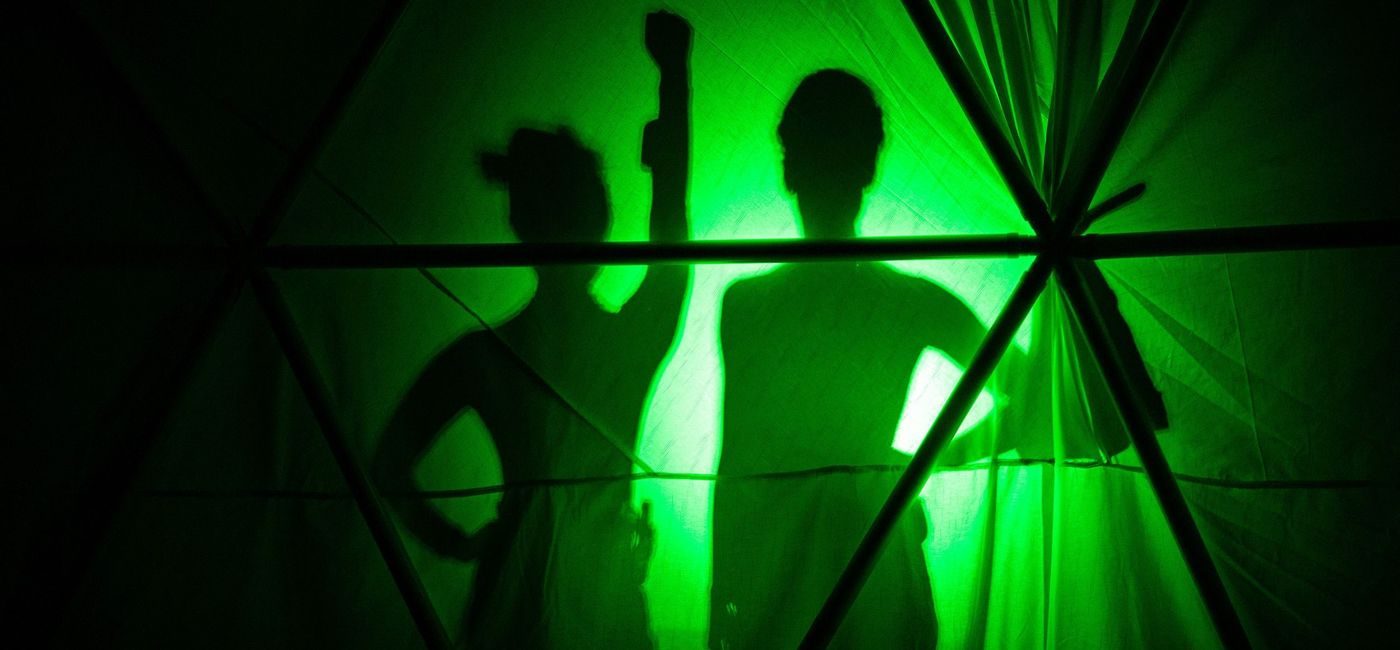 Silhuettene av to kvinnelige utøvere bak et sceneteppe, med grønt lys bak.