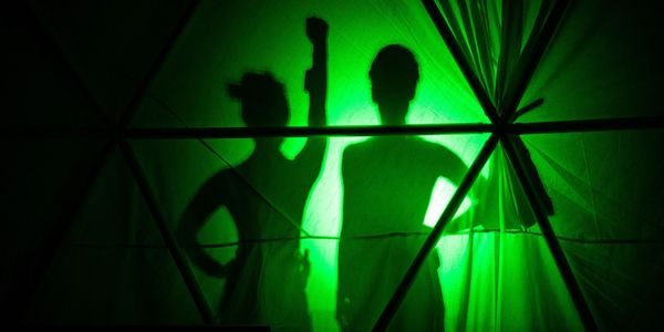 Silhuettene av to kvinnelige utøvere bak et sceneteppe, med grønt lys bak.