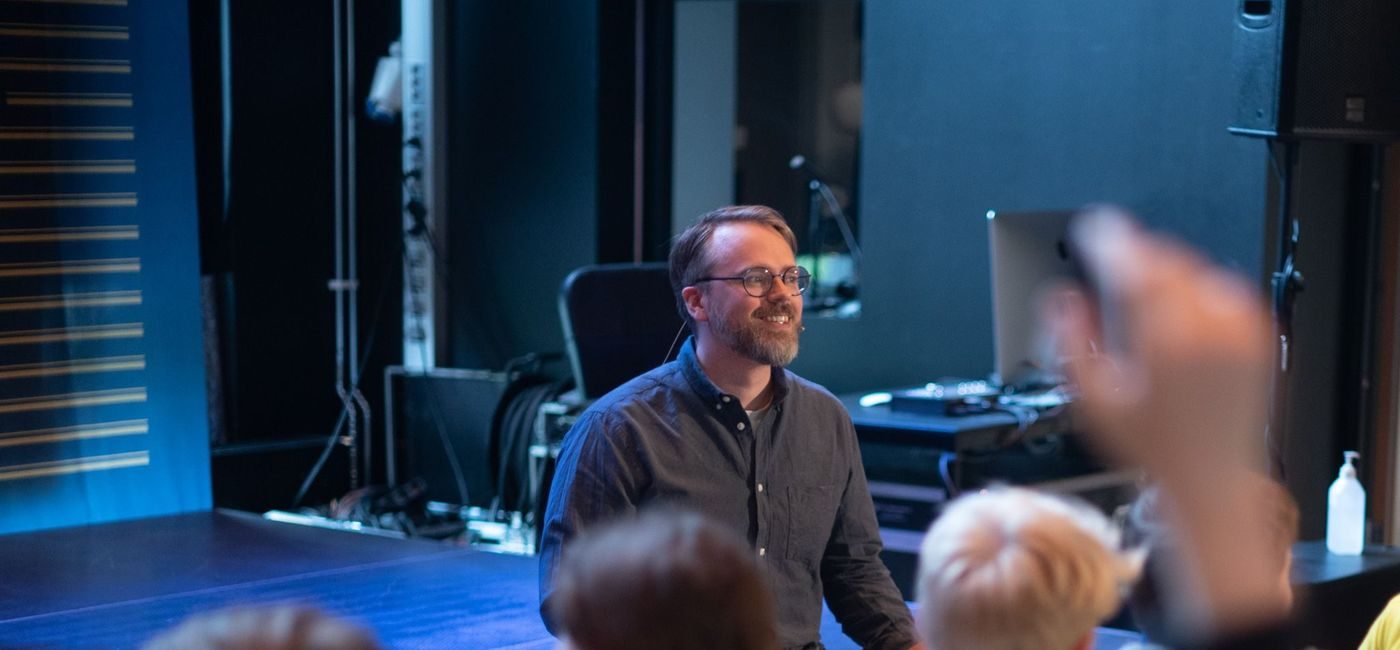 En mann sitter på kanten av en scene og smiler mot sitt publikum, som er flere barn med gule t-skjorter og ryggen mot kamera. En av dem har hånden i været.