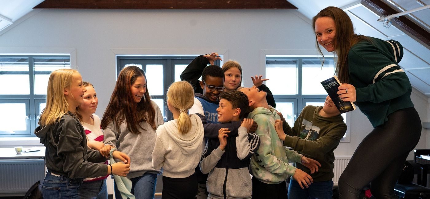 Forfatter Mari Moen Holsve står på en stol foran en gruppe elever som gjør ulike bevegelser og smiler