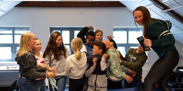 Forfatter Mari Moen Holsve står på en stol foran en gruppe elever som gjør ulike bevegelser og smiler