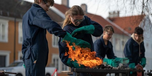 Elever skjermer seg mot flammer som stiger opp mot dem. De har på seg verneutstyr