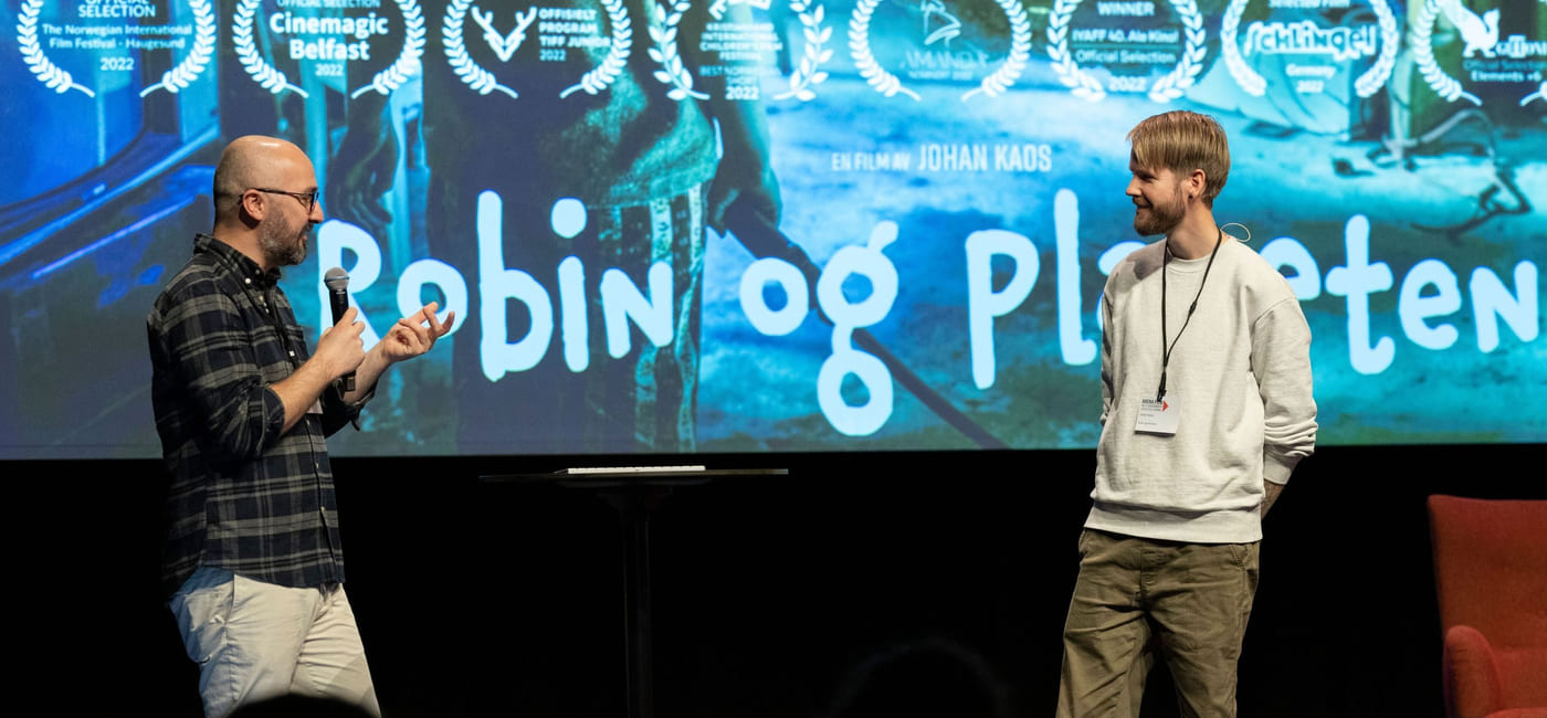 To menn snakker på en scene. Bak dem er det en storskjerm med et bilde av en plakat der det står "Robin og planeten".