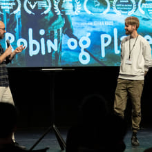 To menn snakker på en scene. Bak dem er det en storskjerm med et bilde av en plakat der det står "Robin og planeten".