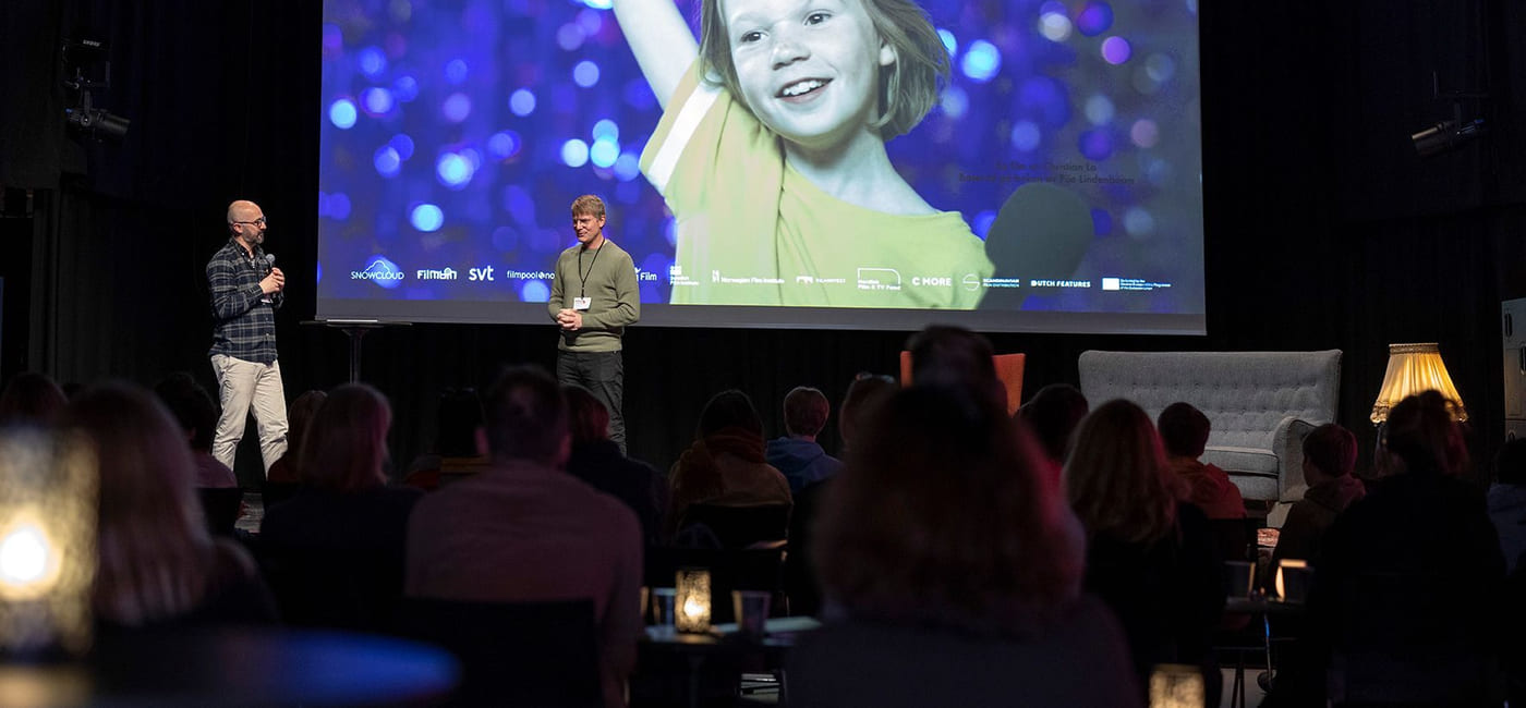 To menn snakker på en scene foran et mørklagt publikum. Bak dem er det en storskjerm med et bilde av en smilende jente som strekker en arm i været.