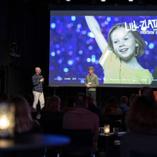 To menn snakker på en scene foran et mørklagt publikum. Bak dem er det en storskjerm med et bilde av en smilende jente som strekker en arm i været.