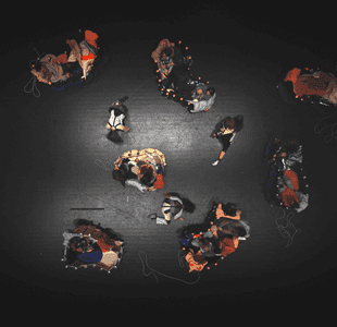 Voksne og barn som sitter i vogner dekket med stoff. Bildet er tatt ovenfra. Vognene står på et svart scenegulv.