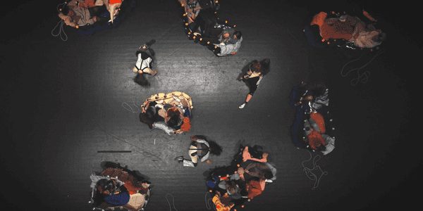 Voksne og barn som sitter i vogner dekket med stoff. Bildet er tatt ovenfra. Vognene står på et svart scenegulv.