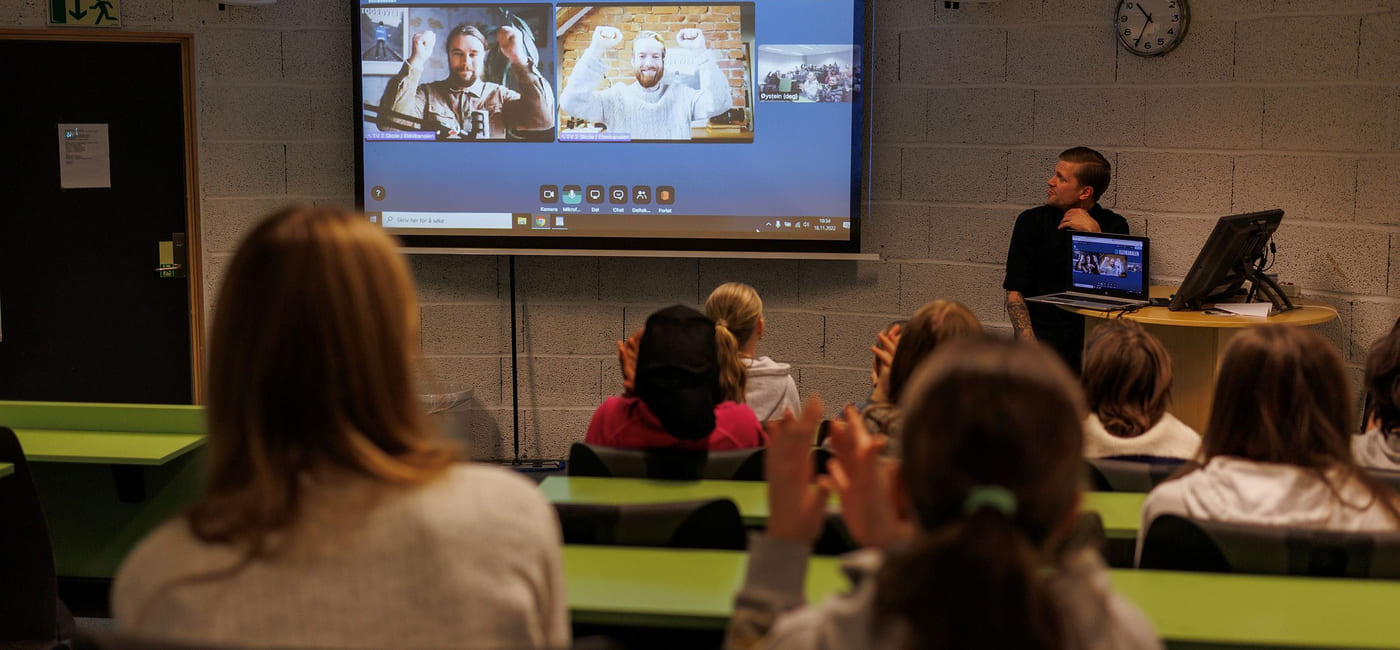 To utøvere i videovinduer på skjerm strekker hendene i været, elever i auditoriet klapper.