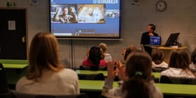 To utøvere i videovinduer på skjerm strekker hendene i været, elever i auditoriet klapper.