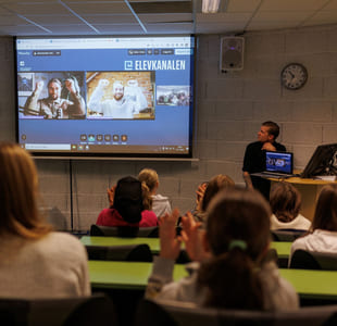 To utøvere i videovinduer på skjerm strekker hendene i været, elever i auditoriet klapper.