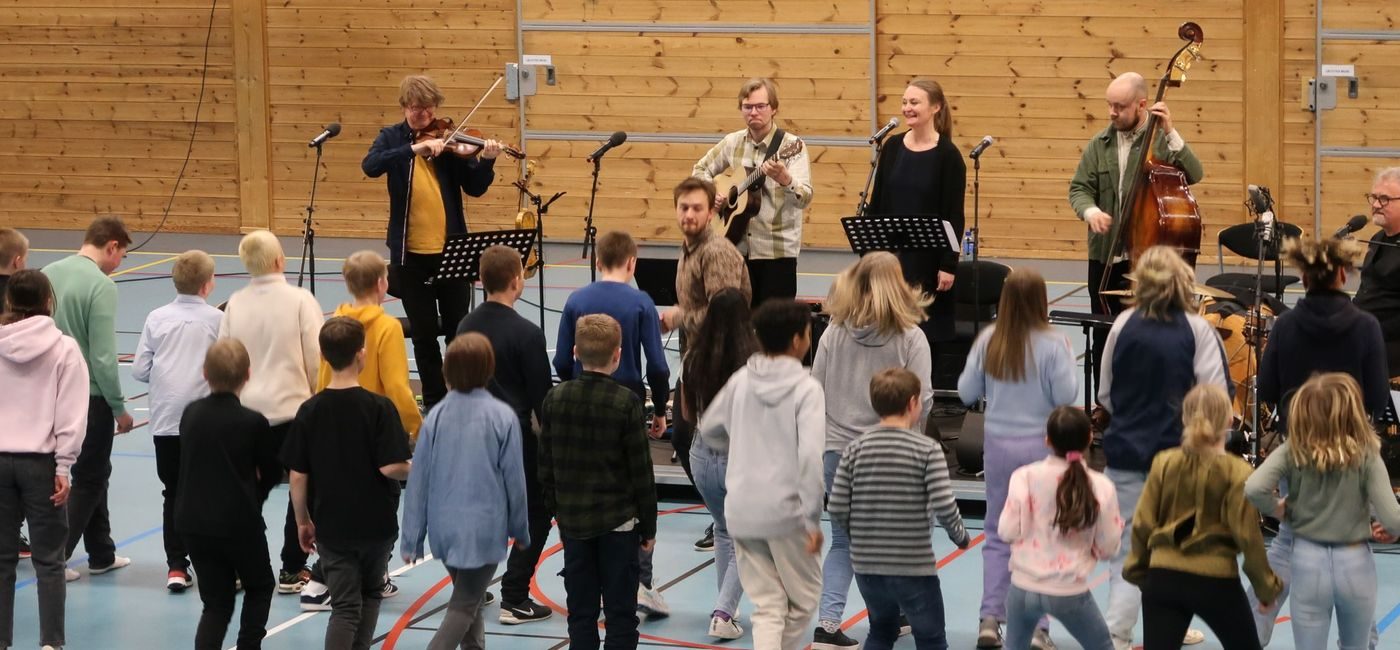 Fem musikere spiller i en gymsal. Barn og unge danser foran dem