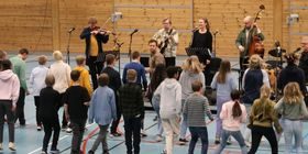 Fem musikere spiller i en gymsal. Barn og unge danser foran dem