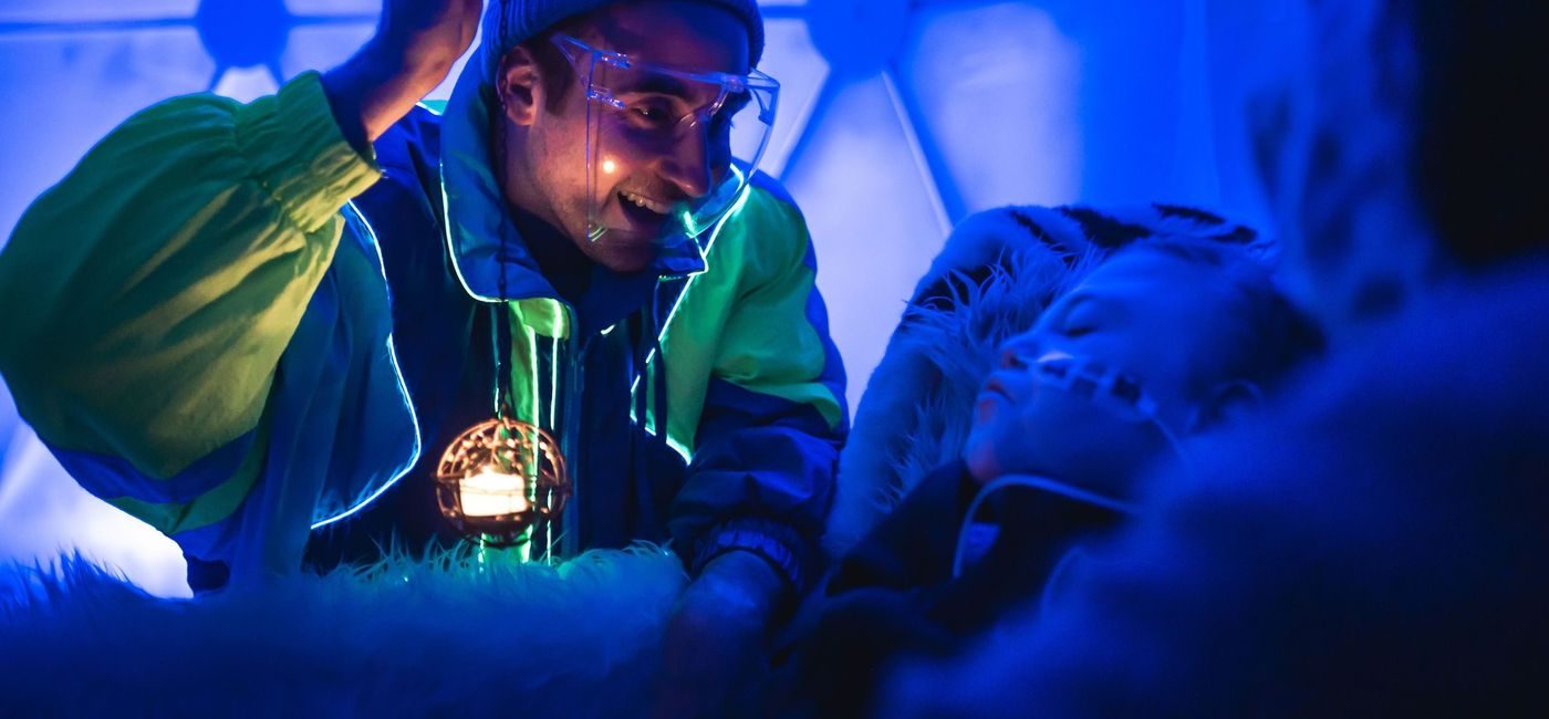 En mann med blå lue og gjennomsiktig skjerm foran ansiktet holder opp en ball med et lys i til et barn med en slange i nesen. De er begge opplyst av blått lys.