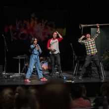 Fire mennesker på en scene. En mann holder en gitar, en annen et slags langt horn som han holder i været, en kvinne holder en mikrofon og peker ut på forsamlingen.