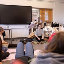 En kvinne sitter på gulvet i et klasserom og snakker til flere ungdommer, de sitter eller ligger på yogamatter.