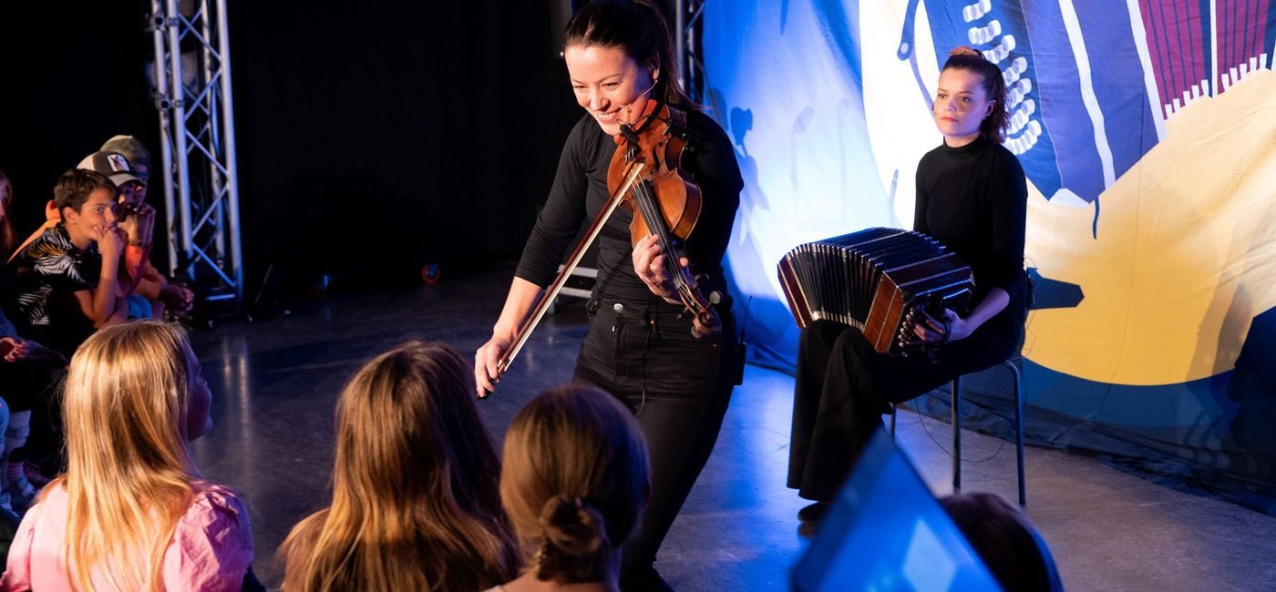 To utøvere med trekkspill og fiolin spiller foran en gruppe elever.