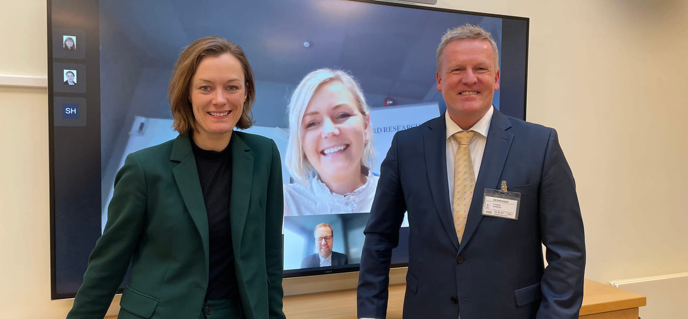 Anette Trettebergstuen og Øystein Strand står foran smilende foran en stor TV-skjerm, der vi kan se en kvinne og en mann som smiler.