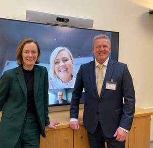 Anette Trettebergstuen og Øystein Strand står foran smilende foran en stor TV-skjerm, der vi kan se en kvinne og en mann som smiler.