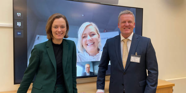 Anette Trettebergstuen og Øystein Strand står foran smilende foran en stor TV-skjerm, der vi kan se en kvinne og en mann som smiler.