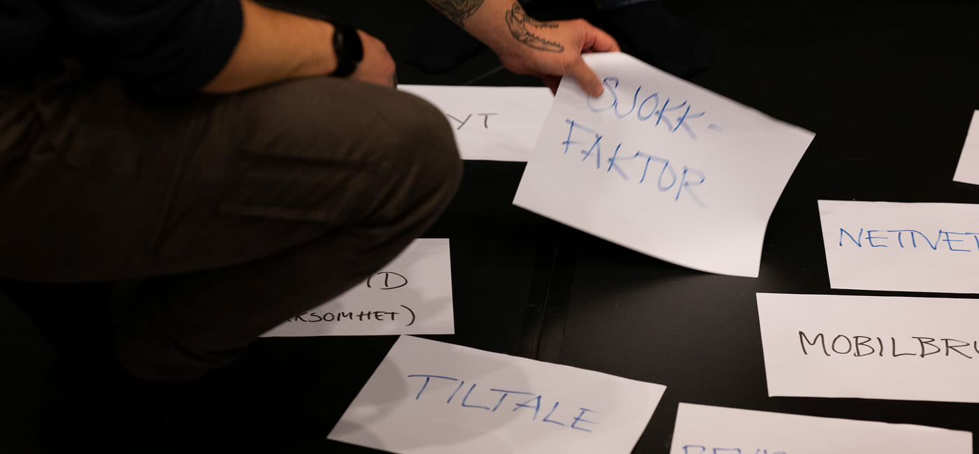 En mann med tatovert hånd lener seg ned og plukker opp et ark med ordet "sjokkfaktor".