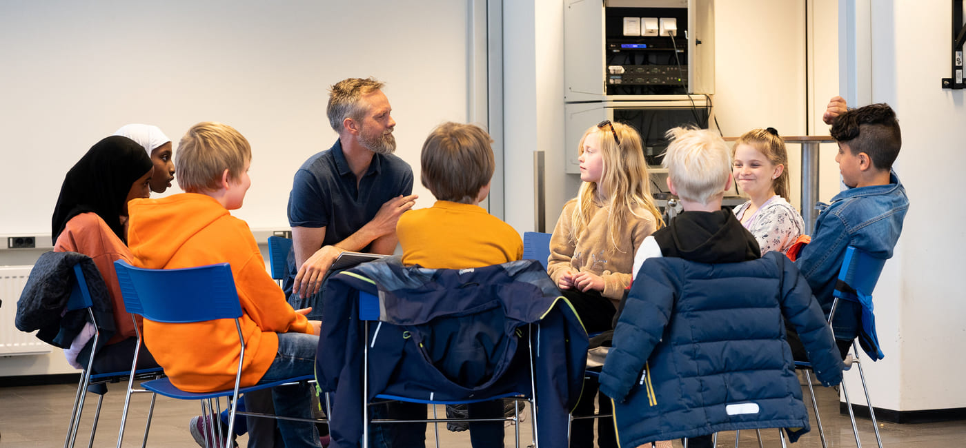 En mann og flere skolebarn sitter på stoler i en sirkel og snakker sammen.