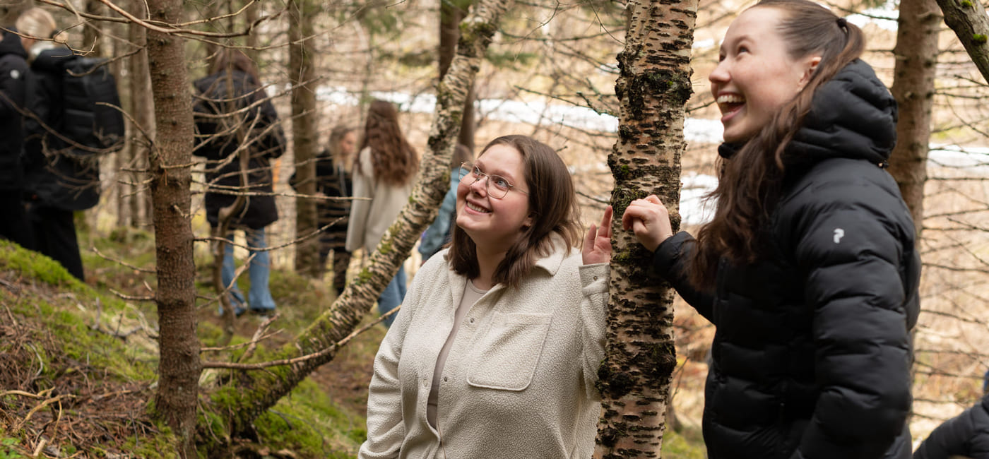 To unge kvinner ser på noe. Begge smiler. De står i en skog.