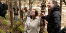 To unge kvinner ser på noe. Begge smiler. De står i en skog.