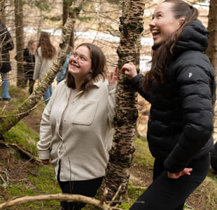 To unge kvinner ser på noe. Begge smiler. De står i en skog.