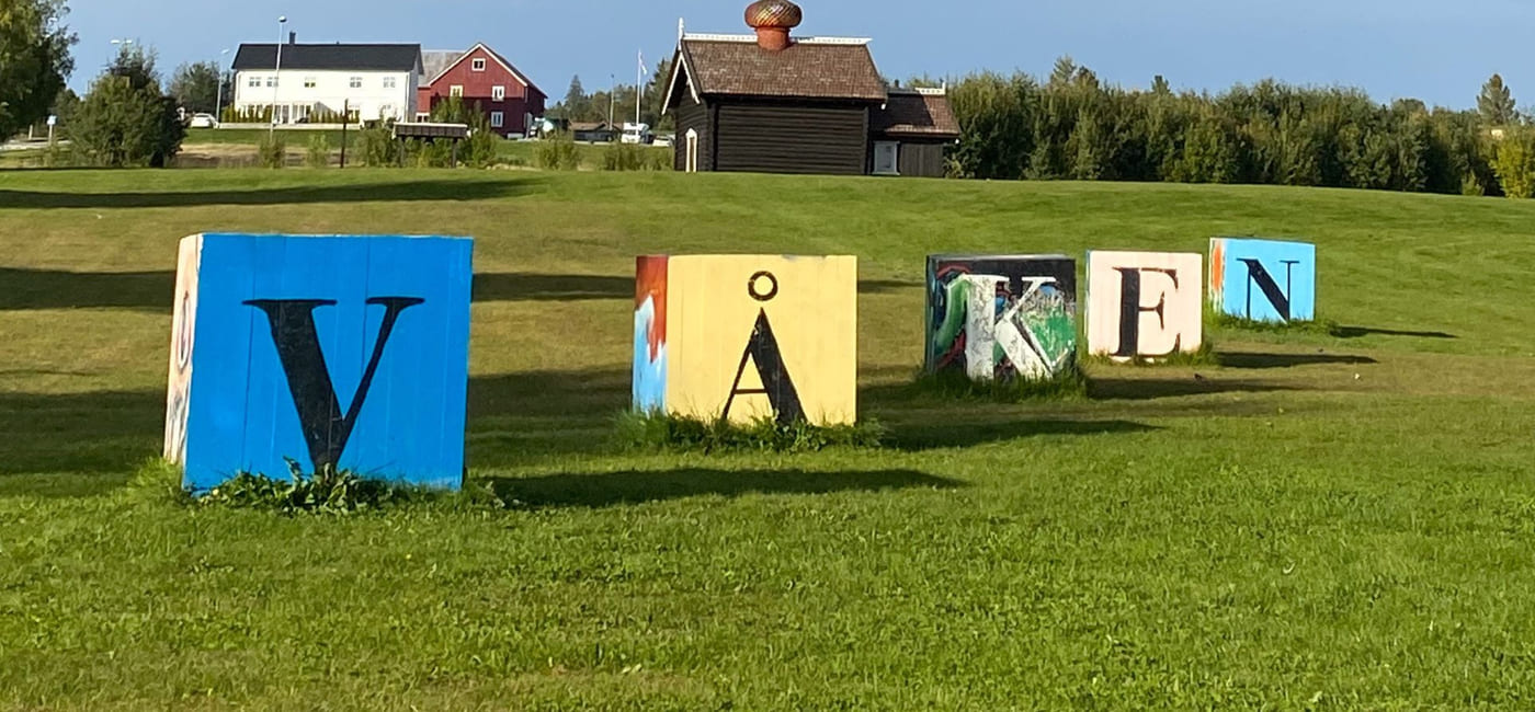 Store, fargerike bokstaver på treklosser som sammen utgjør "Våken". Står på en gressplen foran en gård.
